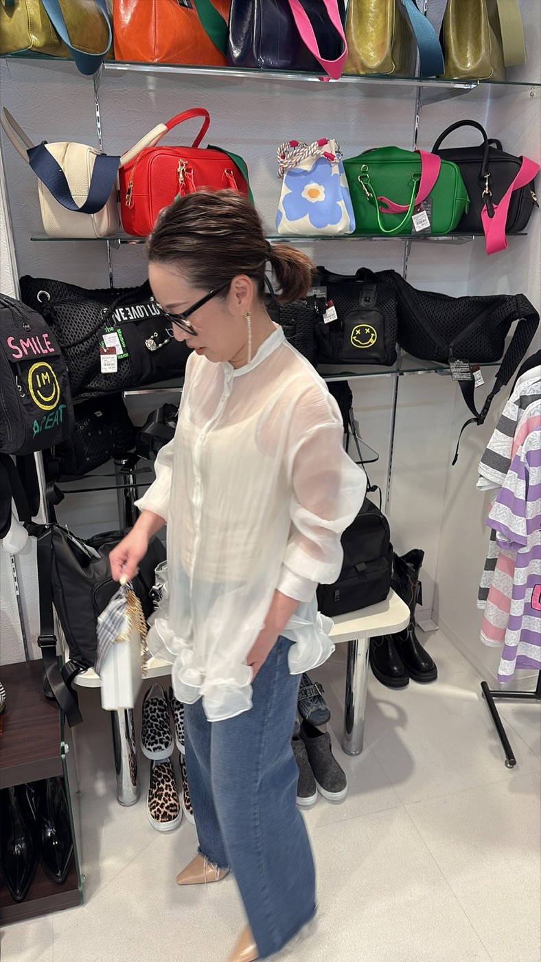 バッグが並ぶ店内で、白いシアーシャツとデニムで小物を手に取る hime